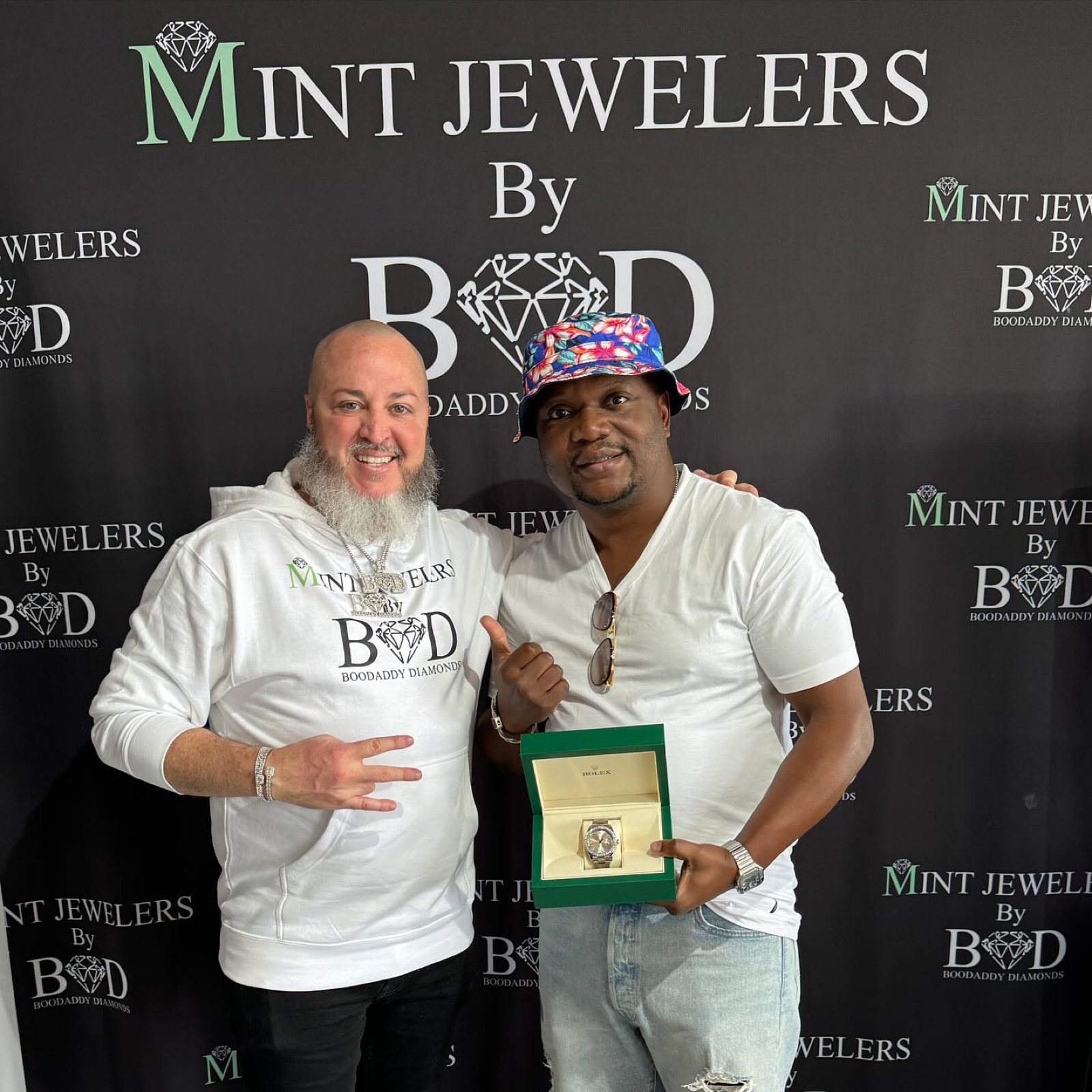 Boodaddy and a customer in front of the Mint Jewelers backdrop, holding a Rolex box
